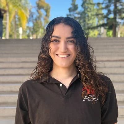 Aita Sharifi, smiling, in a brown ACE Robotics polo on outdoor steps