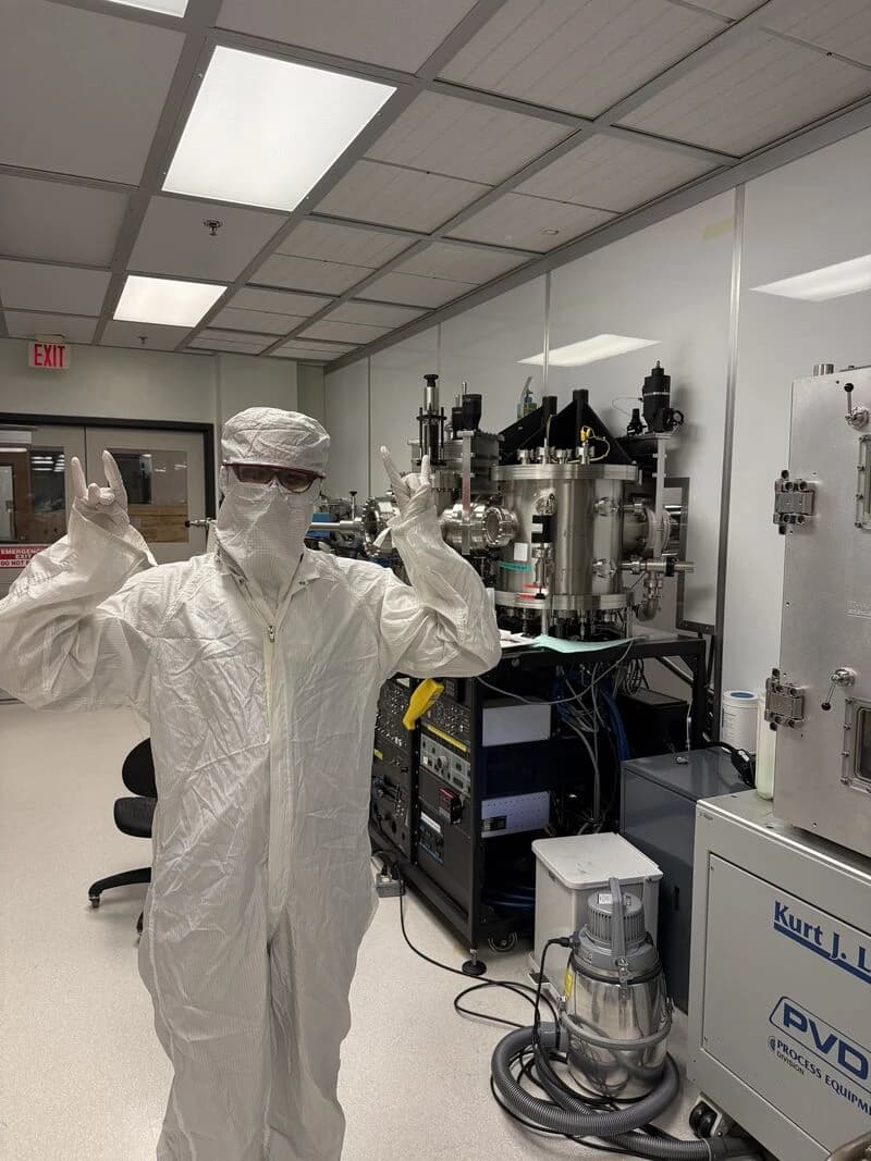 Aita in a full cleanroom gown, hood, mask and safety glasses, next to vacuum deposition equipment