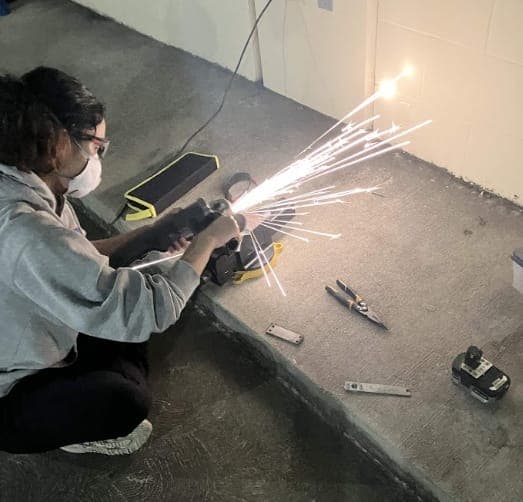 Aita, in a mask and safety glasses, angle grinding the edge of a titanium part; sparks fly toward the wall
